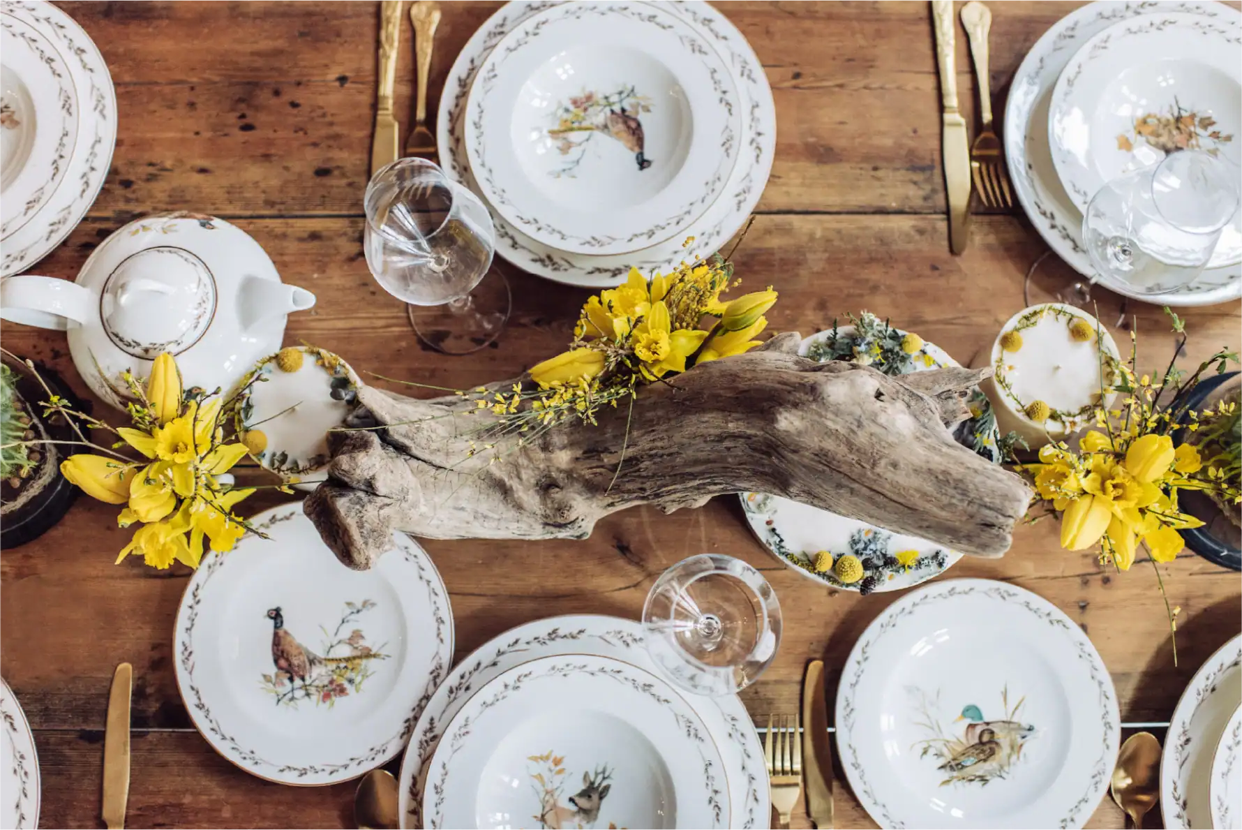 Bella tavola apparecchiata con porcellana floreale, centrotavola in legno e fiori gialli.